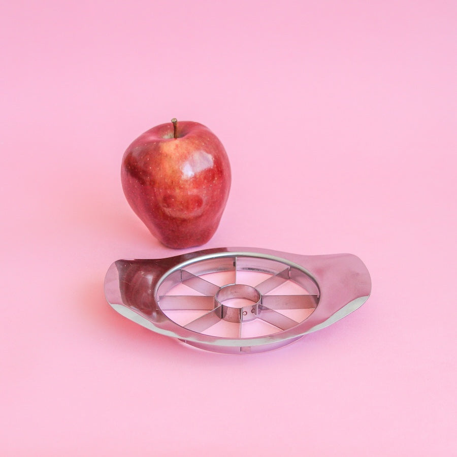 Cortador de manzana de acero junto a una manzana roja sobre fondo rosa
