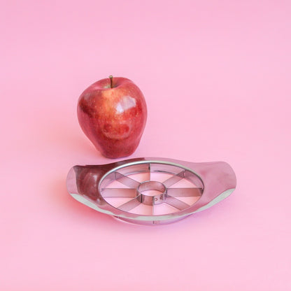 Cortador de manzana de acero junto a una manzana roja sobre fondo rosa
