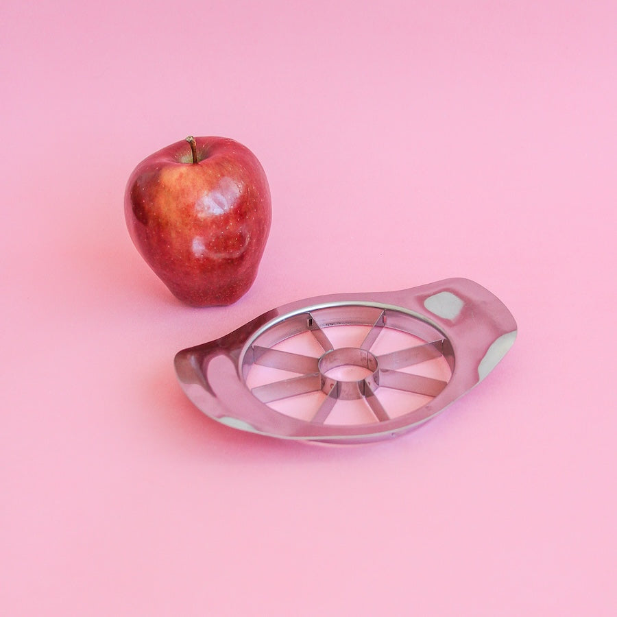Cortador de manzana de acero junto a una manzana roja sobre fondo rosa

