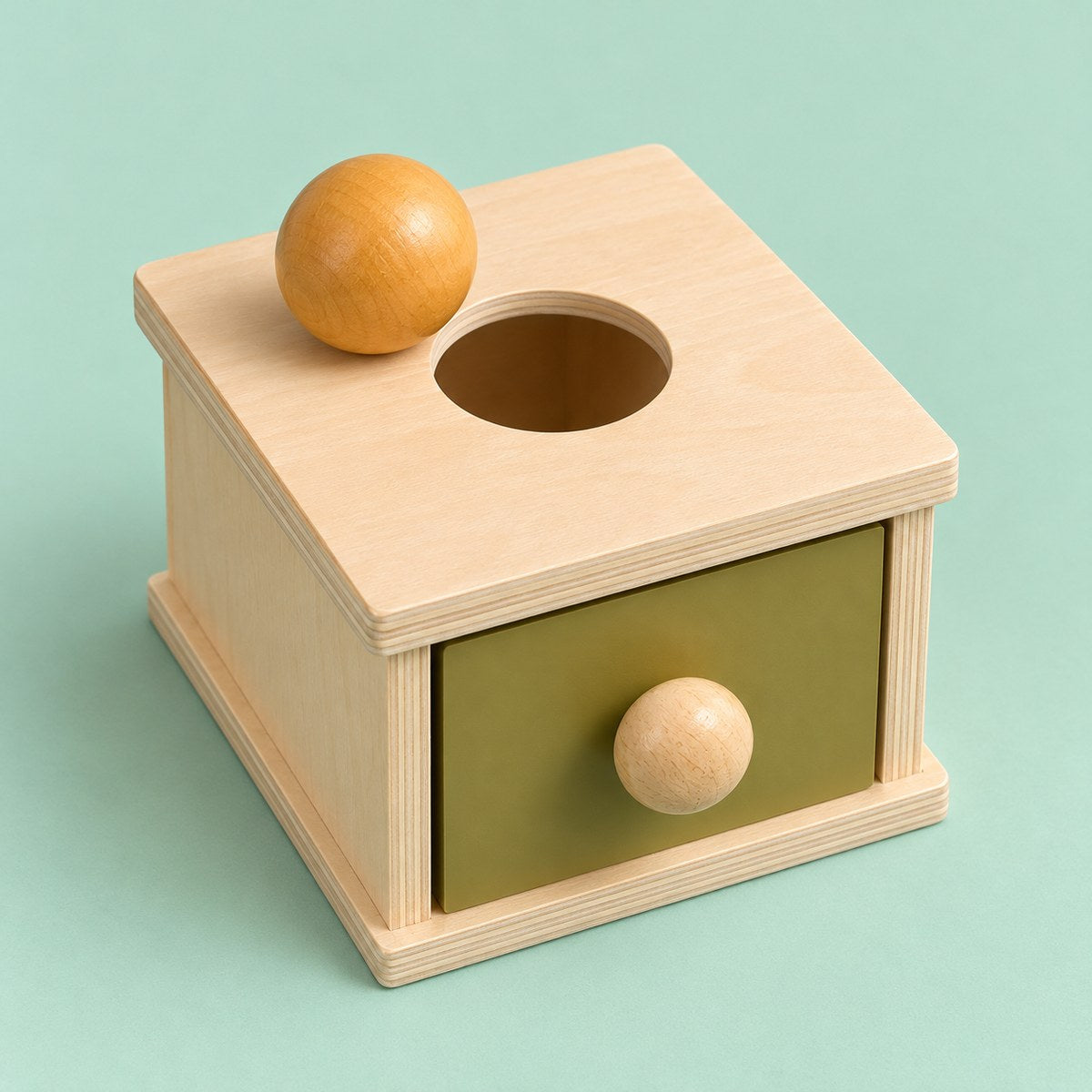 Caja de permanencia Montessori de madera con tiroir verde oliva, pomo de madera y pelota ocre sobre la tapa con agujero